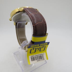 May include: A gold-toned wristwatch with a brown leather band. The watch has a round face and a textured band. A yellow tag with the text "WATER RESIST" and the brand "Q&Q" is attached to the watch.