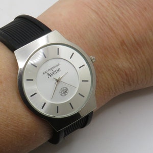 May include: A silver watch with a black silicone band. The watch face is white with black markings and the text "Eau Thermale Av&egrave;ne" printed on it.
