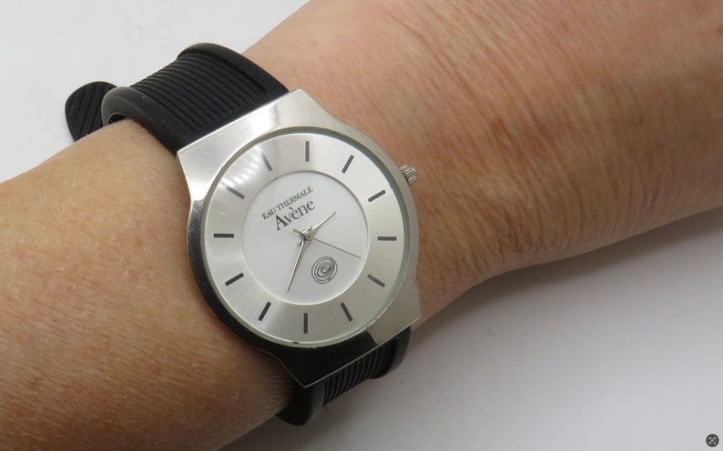 May include: A silver watch with a black silicone band. The watch face is white with black markings and the text "Eau Thermale Av&egrave;ne" printed on it.