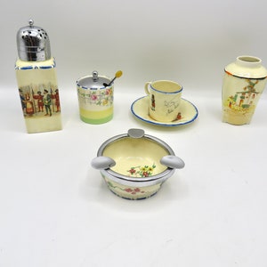 May include: A set of five vintage ceramic pieces with a cream background and colorful floral and figural designs. The set includes a sugar bowl with a spoon, a salt shaker, a small vase, a cup and saucer, and an ashtray with a silver rim.