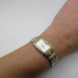 Könnte beinhalten: Eine zweifarbige Silber- und Golduhr mit einem rechteckigen Zifferblatt und einem weißen Zifferblatt. Die Uhr hat ein Citizen-Logo auf dem Zifferblatt.