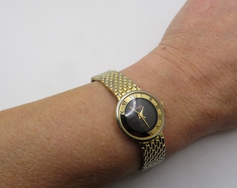 May include: A gold-tone wristwatch with a black face and Roman numeral hour markers. The watch has a gold-tone metal band.