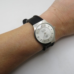 May include: A silver wristwatch with a white face and black rubber band. The watch face has black markings and the text "L'Originale Avenue" printed on it.