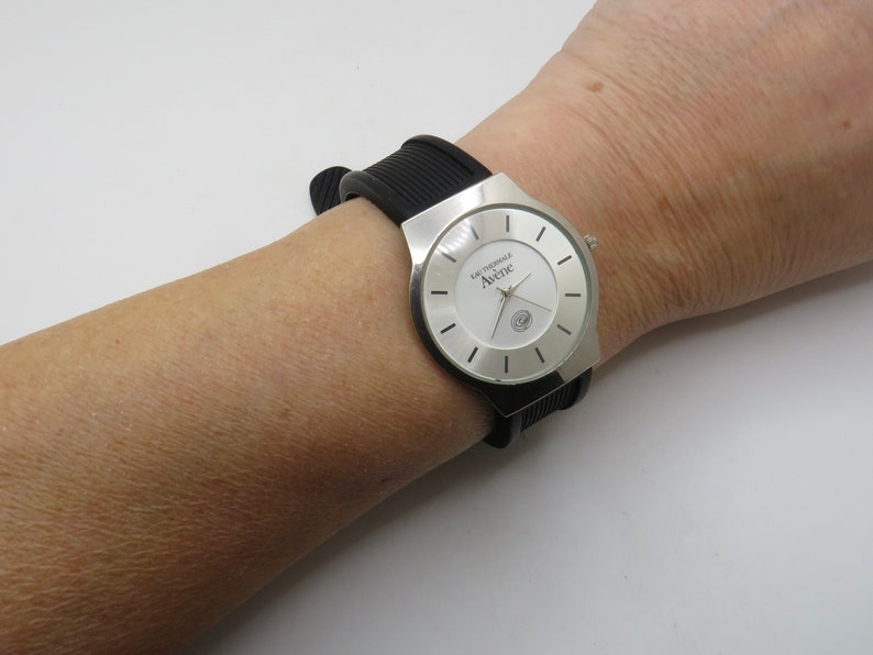 May include: A silver wristwatch with a white face and black rubber band. The watch face has black markings and the text "L'Originale Avenue" printed on it.