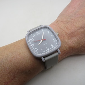May include: A gray quartz watch with a square face and a gray band. The watch face has white numerals and hands, with a red second hand. The watch is on a wrist.