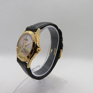 May include: A gold-tone wristwatch with a black leather band. The watch face features a white background with a colorful image of a windmill and the text "Voyage Quartz".