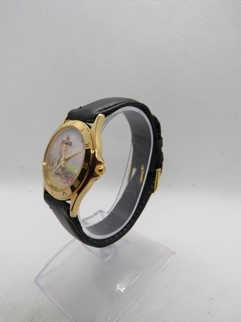May include: A gold-tone wristwatch with a black leather band. The watch face features a white background with a colorful image of a windmill and the text "Voyage Quartz".