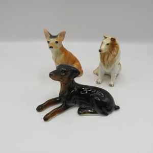 May include: Three ceramic dog figurines. A brown and white chihuahua figurine is standing. A black and brown doberman figurine is lying down. A white and brown collie figurine is sitting.