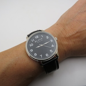 May include: A silver-toned wristwatch with a black leather band and a black face. The watch face has white Arabic numerals and a silver-toned hour and minute hand. The watch face also has the text "BLACK" in white letters.