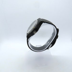 K&ouml;nnte beinhalten: Eine schwarze Armbanduhr mit rundem Zifferblatt und schwarzem Armband. Die Uhr wird auf einem transparenten St&auml;nder vor wei&szlig;em Hintergrund pr&auml;sentiert. Die Uhr hat eine silberfarbene L&uuml;nette und ein schwarzes Zifferblatt.