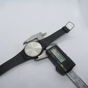 K&ouml;nnte beinhalten: Eine schwarze Armbanduhr mit schwarzem Armband wird mit einem digitalen Messschieber gemessen. Der Messschieber zeigt ein Ma&szlig; von 36,44 mm an. Die Uhr hat einen silberfarbenen R&uuml;cken und ein schwarzes Armband.