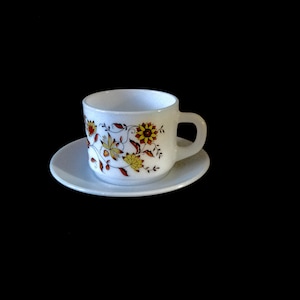 May include: A white teacup and saucer with a floral pattern in shades of yellow, orange, and brown. The cup has a handle and the saucer is round.
