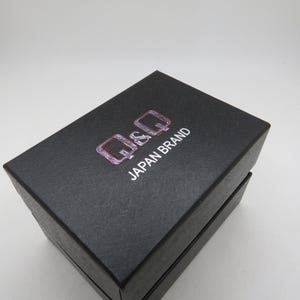May include: A black rectangular box with the text "Q&Q JAPAN BRAND" in white and pink. The box is closed and has a textured surface. The box is likely for a watch or other small item.
