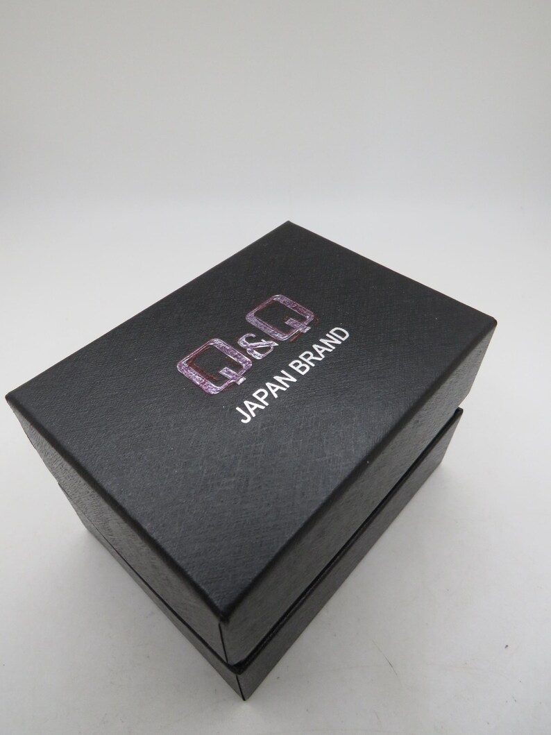 May include: A black rectangular box with the text "Q&Q JAPAN BRAND" in white and pink. The box is closed and has a textured surface. The box is likely for a watch or other small item.