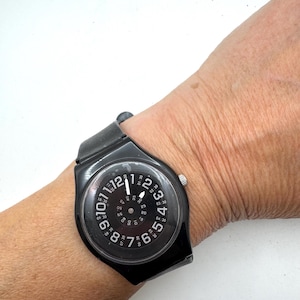 May include: A black wristwatch featuring a round dial with large white Arabic numerals from 1 to 12 around the outer edge and a smaller inner ring of numbers from 05 to 60, with a black strap and a silver-toned crown on the side of the case.