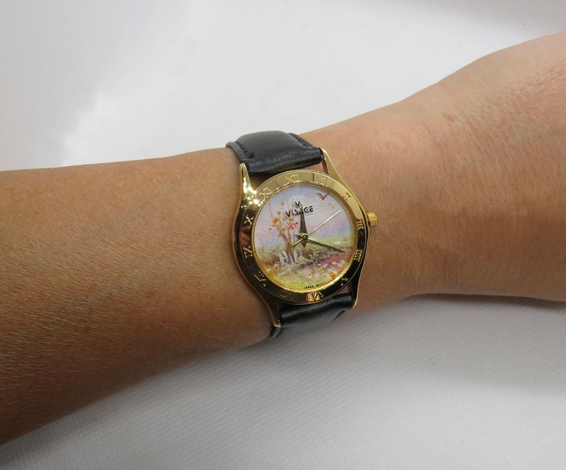 May include: A gold-tone wristwatch with a black leather band. The watch face features a painted scene of a landscape with a tower and trees. The watch face also has Roman numeral hour markers and the word "VISAGE" printed on it.