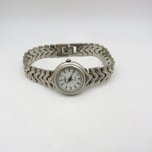 K&ouml;nnte beinhalten: Eine silberne Armbanduhr mit wei&szlig;em Zifferblatt und einem silbernen Metallband. Das Band hat ein einzigartiges Chevron-Muster.