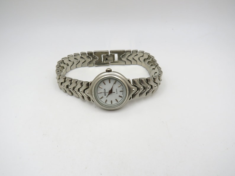 K&ouml;nnte beinhalten: Eine silberne Armbanduhr mit wei&szlig;em Zifferblatt und einem silbernen Metallband. Das Band hat ein einzigartiges Chevron-Muster.