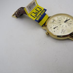 May include: A gold-toned Q&Q watch with a cream-colored face and brown leather band. The watch features two sub-dials and the text "Dual Time" and "Water Resist". The watch is new with a yellow and blue tag.