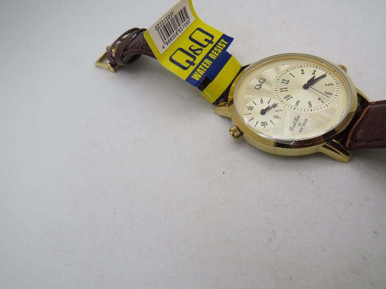May include: A gold-toned Q&Q watch with a cream-colored face and brown leather band. The watch features two sub-dials and the text "Dual Time" and "Water Resist". The watch is new with a yellow and blue tag.