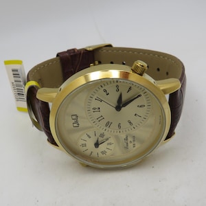 May include: A gold-toned wristwatch with a brown leather band. The watch face has a cream-colored background with black numerals and hands. It features two smaller dials and a crown. The band is a dark brown color.