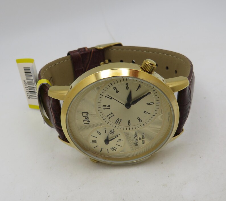 May include: A gold-toned wristwatch with a brown leather band. The watch face has a cream-colored background with black numerals and hands. It features two smaller dials and a crown. The band is a dark brown color.