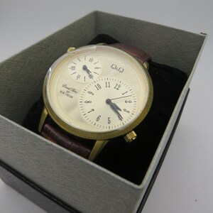 May include: A gold-toned wristwatch with a cream-colored face and three sub-dials, set in a gray box. The watch features a brown leather band. The watch face displays the text "Dual Time" and "WR 5BAR".
