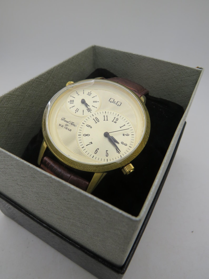 May include: A gold-toned wristwatch with a cream-colored face and three sub-dials, set in a gray box. The watch features a brown leather band. The watch face displays the text "Dual Time" and "WR 5BAR".
