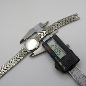 K&ouml;nnte beinhalten: Eine silberne Uhr mit einem Metallband wird mit einer digitalen Schieblehre gemessen. Die Schieblehre zeigt 24,05 mm an.