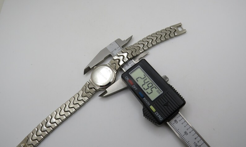 K&ouml;nnte beinhalten: Eine silberne Uhr mit einem Metallband wird mit einer digitalen Schieblehre gemessen. Die Schieblehre zeigt 24,05 mm an.