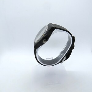 K&ouml;nnte beinhalten: Eine schwarze Armbanduhr mit rundem Zifferblatt und schwarzem Armband. Die Uhr wird auf einem transparenten St&auml;nder vor wei&szlig;em Hintergrund pr&auml;sentiert. Die Uhr hat eine silberfarbene L&uuml;nette und ein schwarzes Zifferblatt.