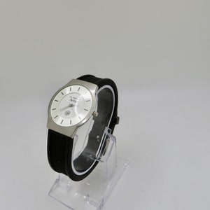 May include: A silver watch with a white face and black rubber band. The watch face has the text "La Cosmopolite" and "Aurea" printed on it.