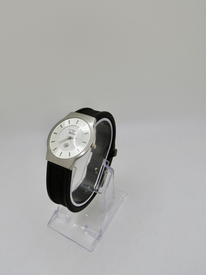 May include: A silver watch with a white face and black rubber band. The watch face has the text "La Cosmopolite" and "Aurea" printed on it.