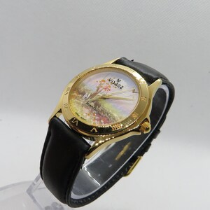 May include: A gold-tone wristwatch with a black leather band. The watch face features a white background with a painted landscape scene and the word "Village" in gold lettering. The watch face also has Roman numeral hour markers.