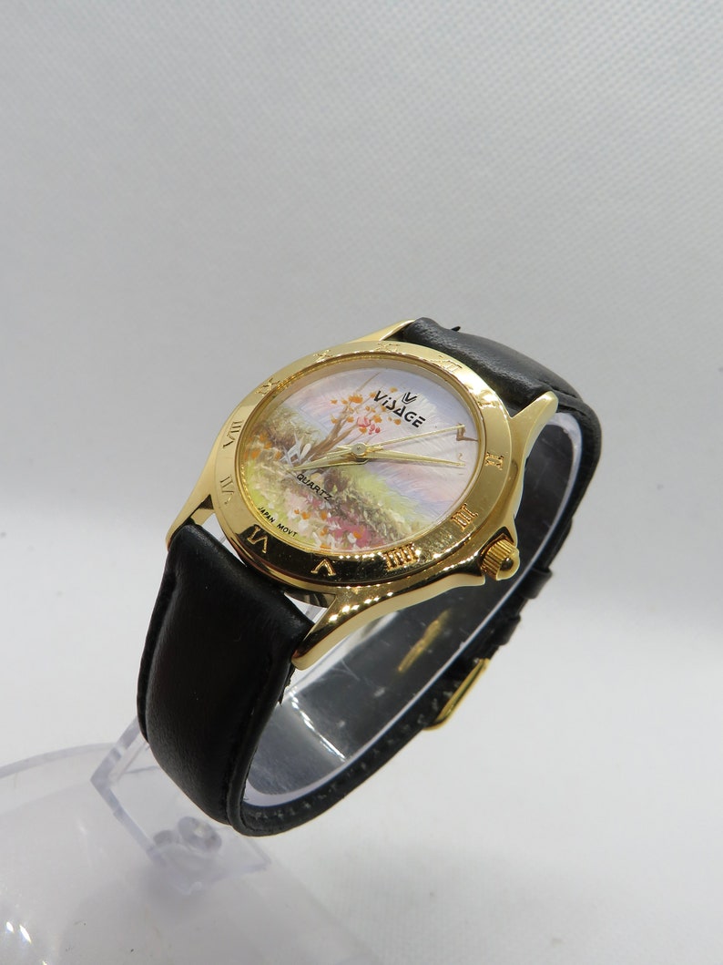 May include: A gold-tone wristwatch with a black leather band. The watch face features a white background with a painted landscape scene and the word "Village" in gold lettering. The watch face also has Roman numeral hour markers.