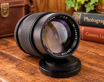 May include: A black Unitor Auto 135mm f/2.8 camera lens with a textured focus ring and a serial number 164879 printed on the front element rim. The lens is positioned horizontally with a matching black circular lens cap resting directly in front of it.