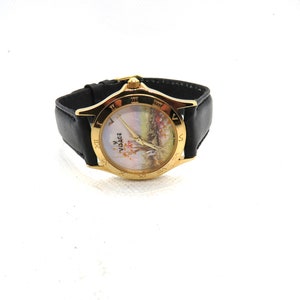 May include: A gold-tone Versace watch with a black leather band. The watch face features a white background with a floral design and Roman numeral hour markers.