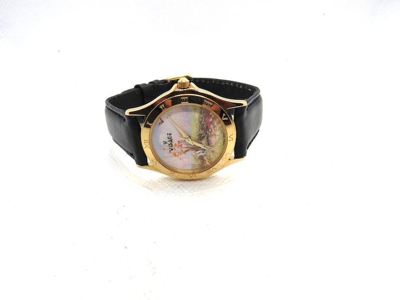 May include: A gold-tone Versace watch with a black leather band. The watch face features a white background with a floral design and Roman numeral hour markers.