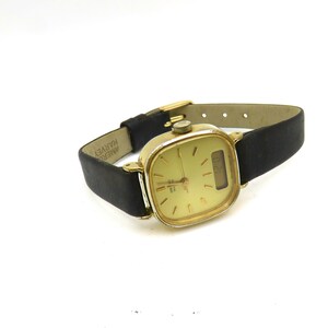 K&ouml;nnte beinhalten: Eine goldfarbene analoge Armbanduhr mit quadratischem Zifferblatt und schwarzem Lederarmband. Das Zifferblatt hat einen wei&szlig;en Hintergrund mit schwarzen Zeigern und einem Datumsfenster. Das Armband ist mit "AMER. HARVEY" beschriftet.
