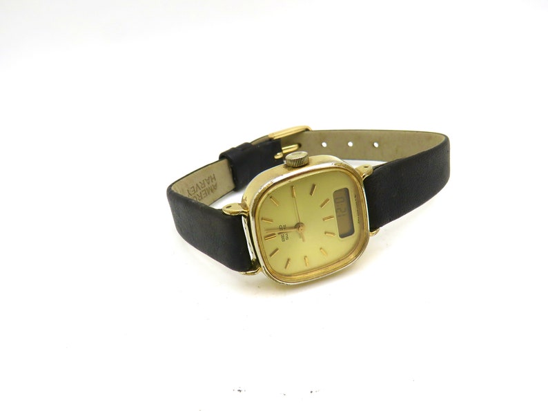 K&ouml;nnte beinhalten: Eine goldfarbene analoge Armbanduhr mit quadratischem Zifferblatt und schwarzem Lederarmband. Das Zifferblatt hat einen wei&szlig;en Hintergrund mit schwarzen Zeigern und einem Datumsfenster. Das Armband ist mit "AMER. HARVEY" beschriftet.