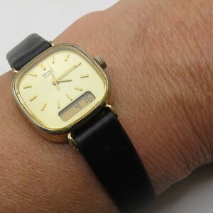 K&ouml;nnte beinhalten: Eine goldfarbene Armbanduhr mit quadratischem Zifferblatt und schwarzem Lederarmband. Die Uhr hat ein digitales Display, das die Zeit 4:16 anzeigt.