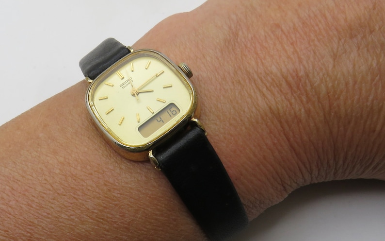 K&ouml;nnte beinhalten: Eine goldfarbene Armbanduhr mit quadratischem Zifferblatt und schwarzem Lederarmband. Die Uhr hat ein digitales Display, das die Zeit 4:16 anzeigt.