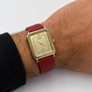May include: A rectangular Citizen Quartz watch with a gold-tone dial, thin gold hands, and the word ALARM printed on the face. The watch features a silver-tone metal bezel and a textured red leather strap with a visible grain pattern.