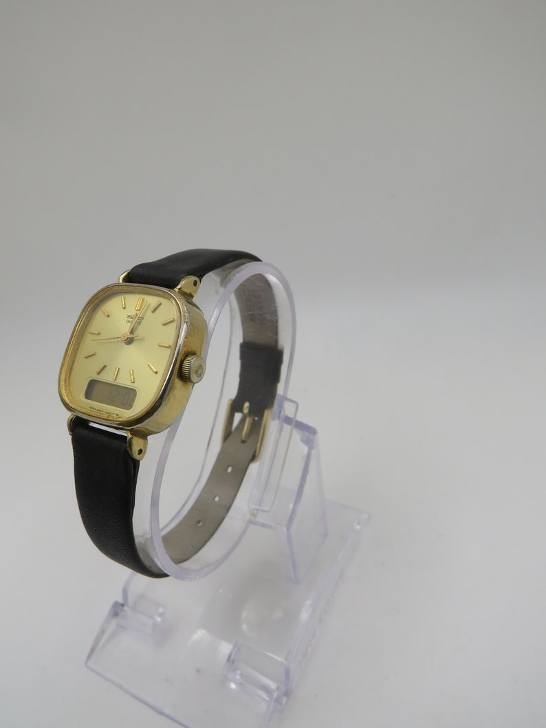 K&ouml;nnte beinhalten: Eine goldfarbene Armbanduhr mit quadratischem Zifferblatt und schwarzem Lederarmband. Die Uhr verf&uuml;gt &uuml;ber ein digitales Display und analoge Zeiger.