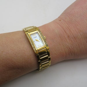 Könnte beinhalten: Eine goldfarbene Uhr mit einem rechteckigen Zifferblatt und einem weißen Zifferblatt. Die Uhr hat eine mit Diamanten besetzte Lünette und ein goldfarbenes Metallband.
