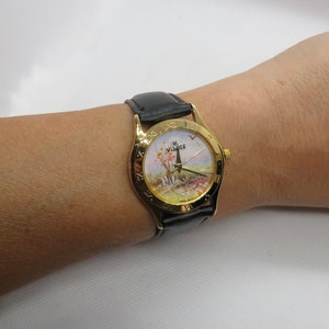 May include: A gold-tone wristwatch with a black leather band. The watch face features a painted scene of a landscape with a tower and trees. The watch face also has Roman numeral hour markers and the word "VISAGE" printed on it.