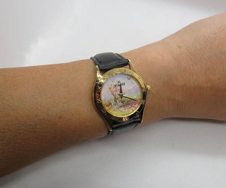 May include: A gold-tone wristwatch with a black leather band. The watch face features a painted scene of a landscape with a tower and trees. The watch face also has Roman numeral hour markers and the word "VISAGE" printed on it.