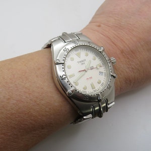 May include: A silver Tissot 1853 PR 200 wristwatch with a white face and a date window. The watch has a silver band and a black bezel.