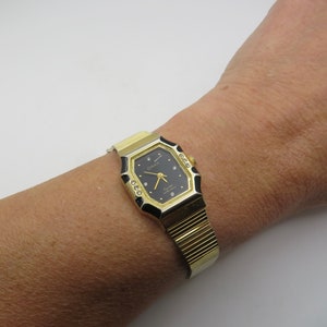 May include: A gold-tone watch with a black face and diamond accents. The watch has a rectangular shape with rounded corners and a gold-tone metal band.