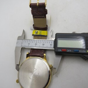 May include: A gold-toned wristwatch with a brown leather band is measured with a digital caliper. The watch face is visible, and the band is partially unclasped. The caliper displays a measurement in millimeters and inches.